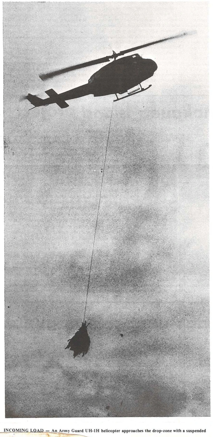 An Army National Guard UH-1H helicopter approaching a drop-zone with a bundle of confiscated cannabis suspended on a long line beneath it, Hawaii, 1979
